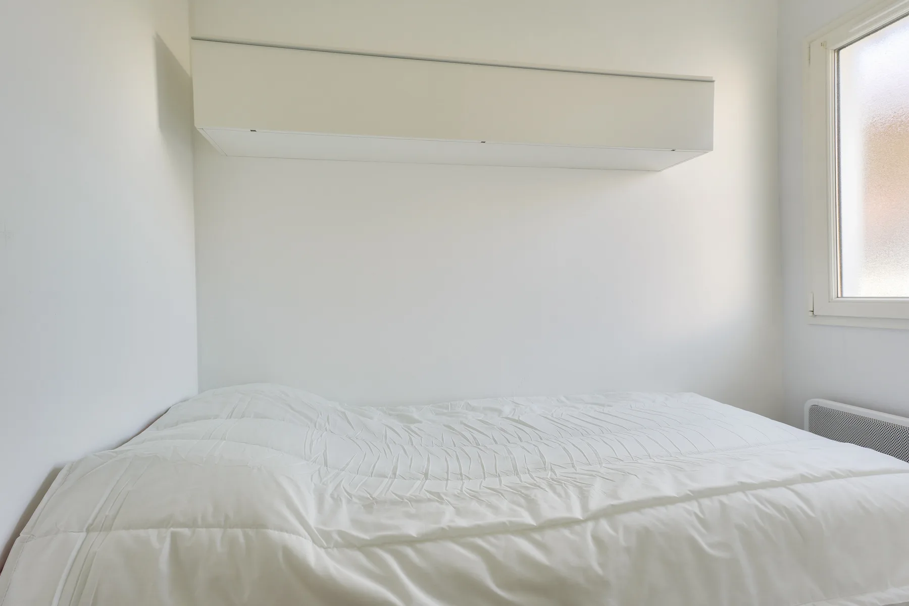 A bedroom with a bed beneath a wall-mounted shelf and a window.