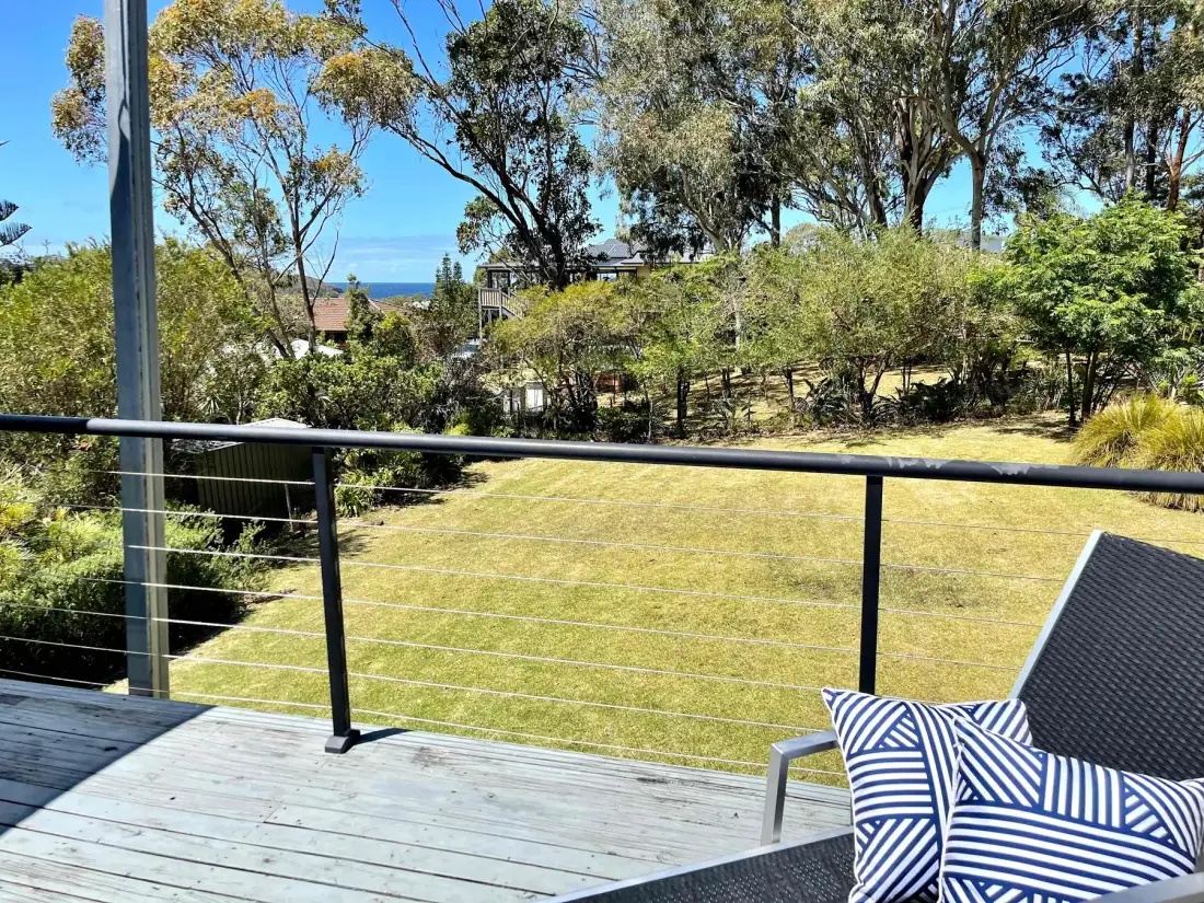 A deck overlooks a grassy yard, trees, and a glimpse of the sea.