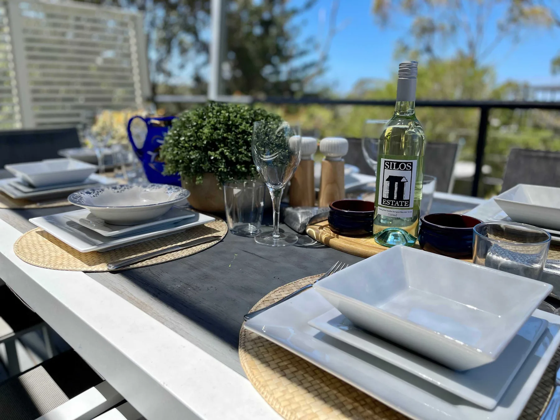 An outdoor dining table is set with plates, glasses, and a bottle.
