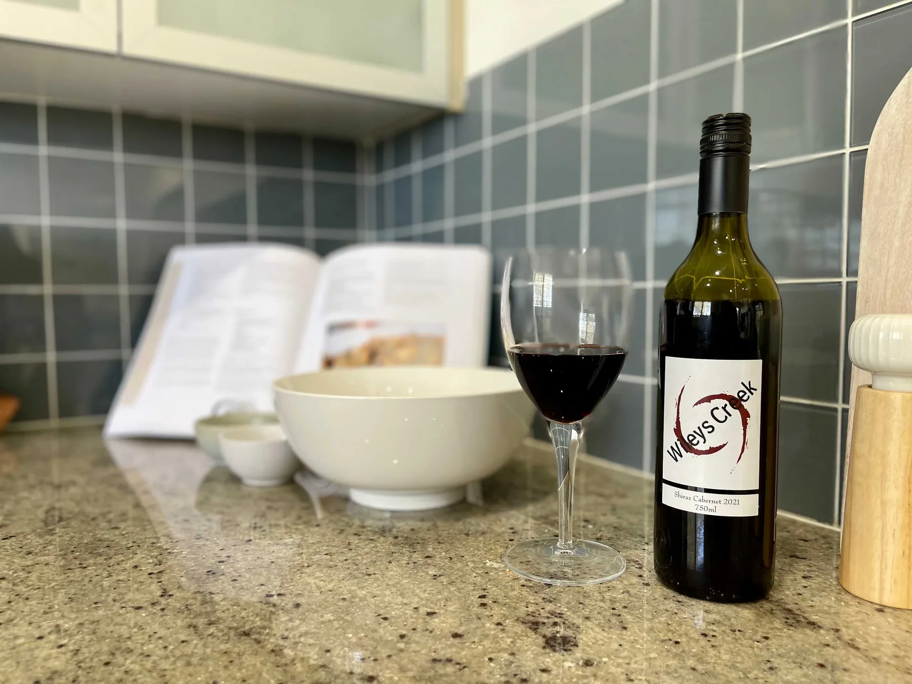 A kitchen counter displays a wine bottle, glass, and bowl against a tiled backsplash.
