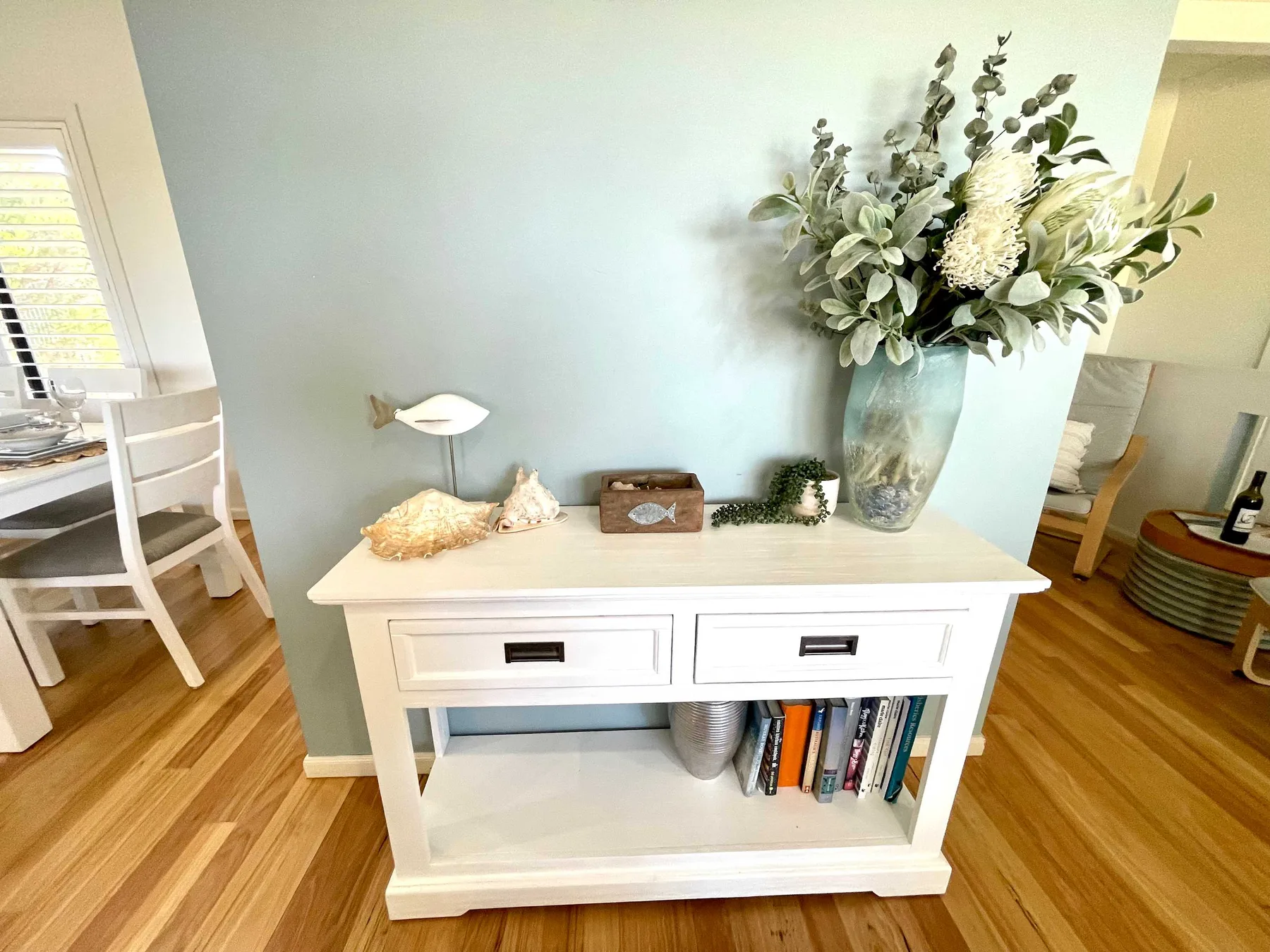 A white console table with books and a large floral arrangement stands against a pale blue wall.
