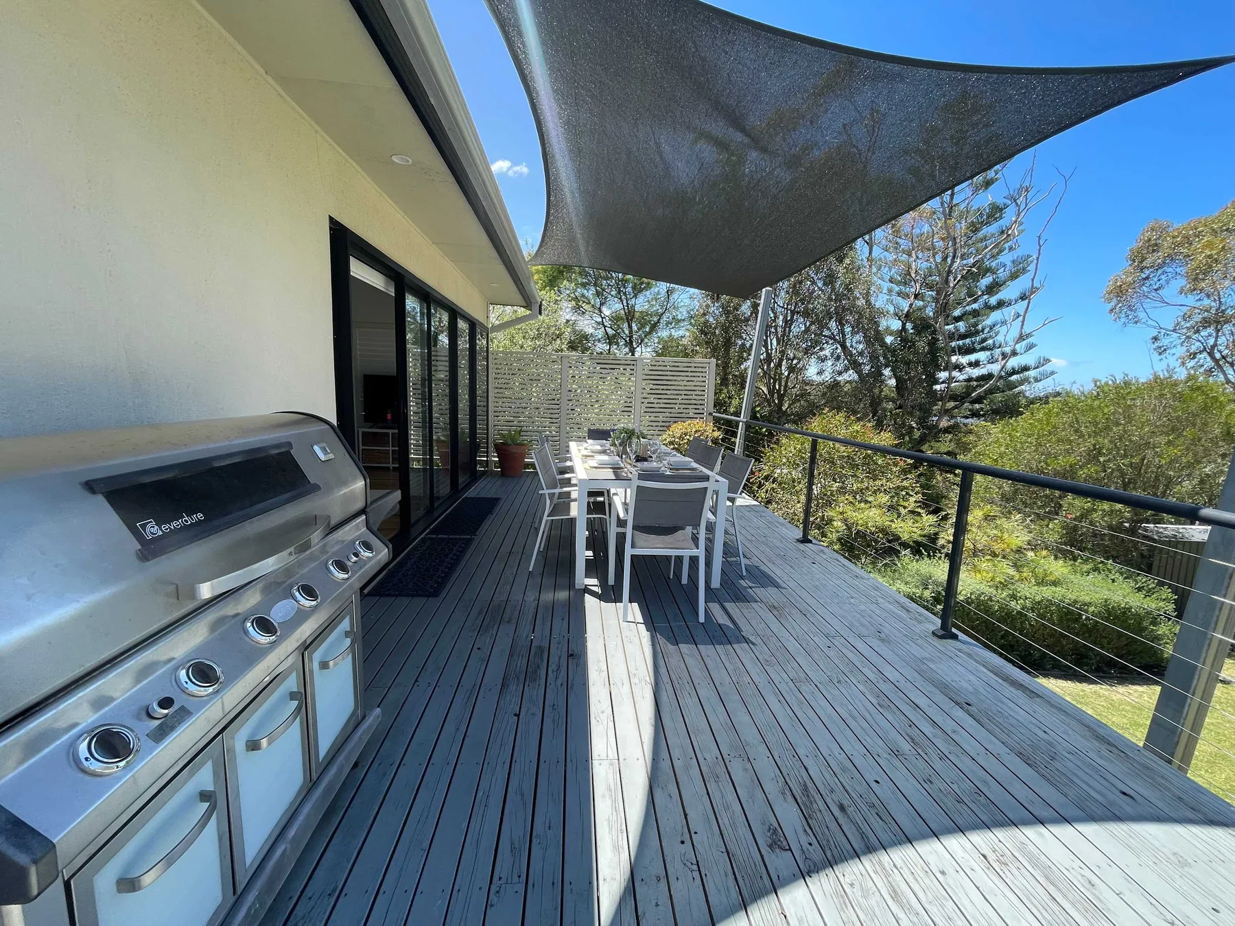 An outdoor deck has a barbecue, dining table, and overhead shade sail.