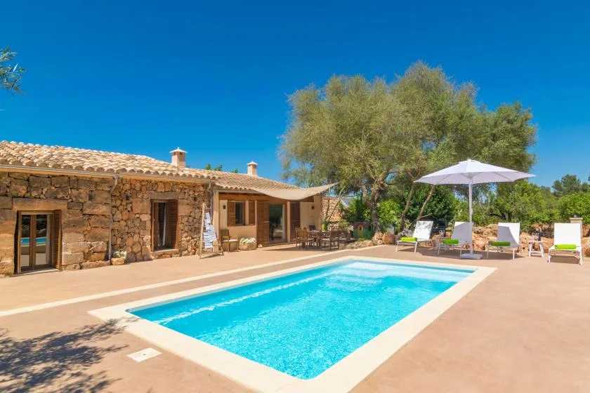 Una piscina y asientos al aire libre junto a una casa de piedra bajo un cielo azul despejado.