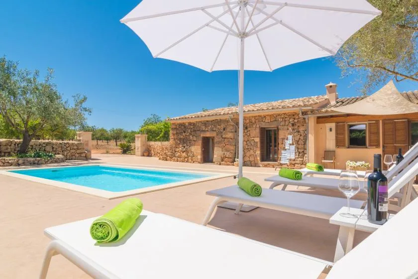 Una casa de piedra da a una piscina, con tumbonas bajo una gran sombrilla blanca.