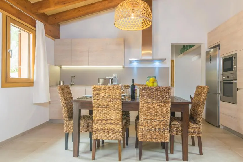 Comedor con mesa de madera, asientos tejidos y una cocina al fondo.