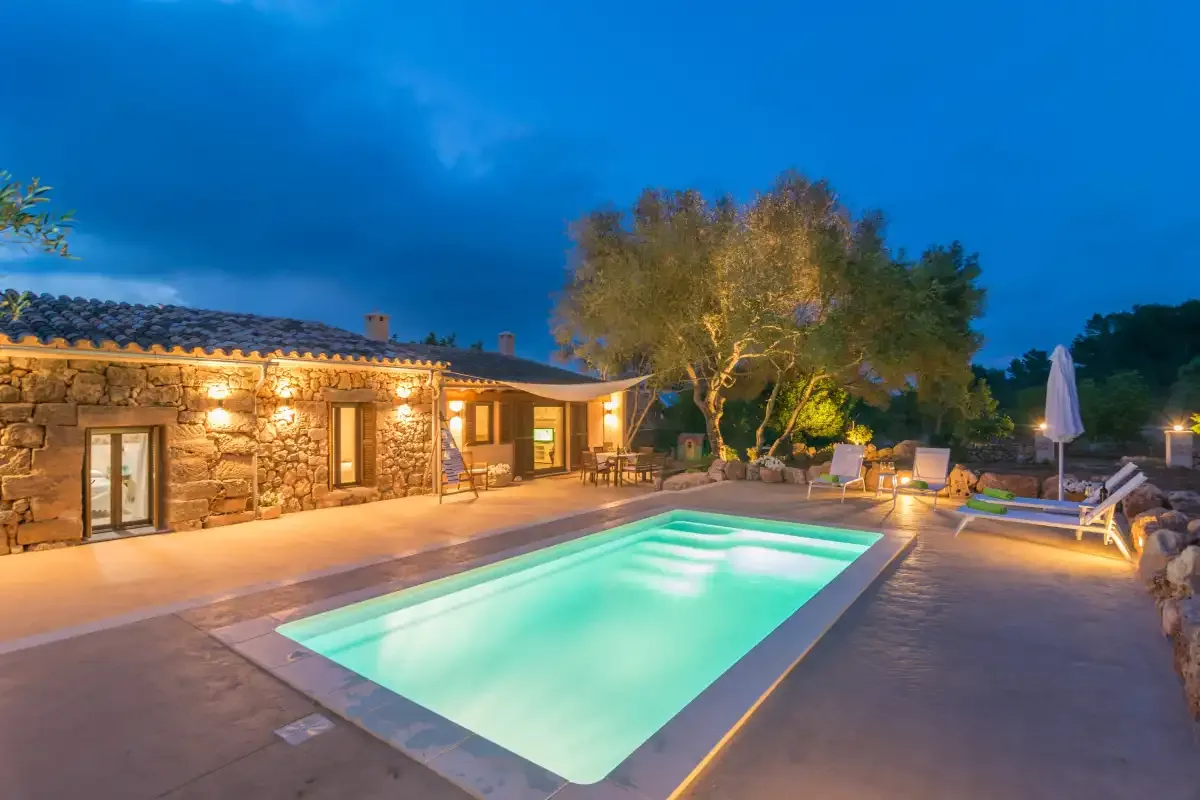 Una casa de piedra y una piscina iluminada junto a una zona de estar al aire libre al anochecer.