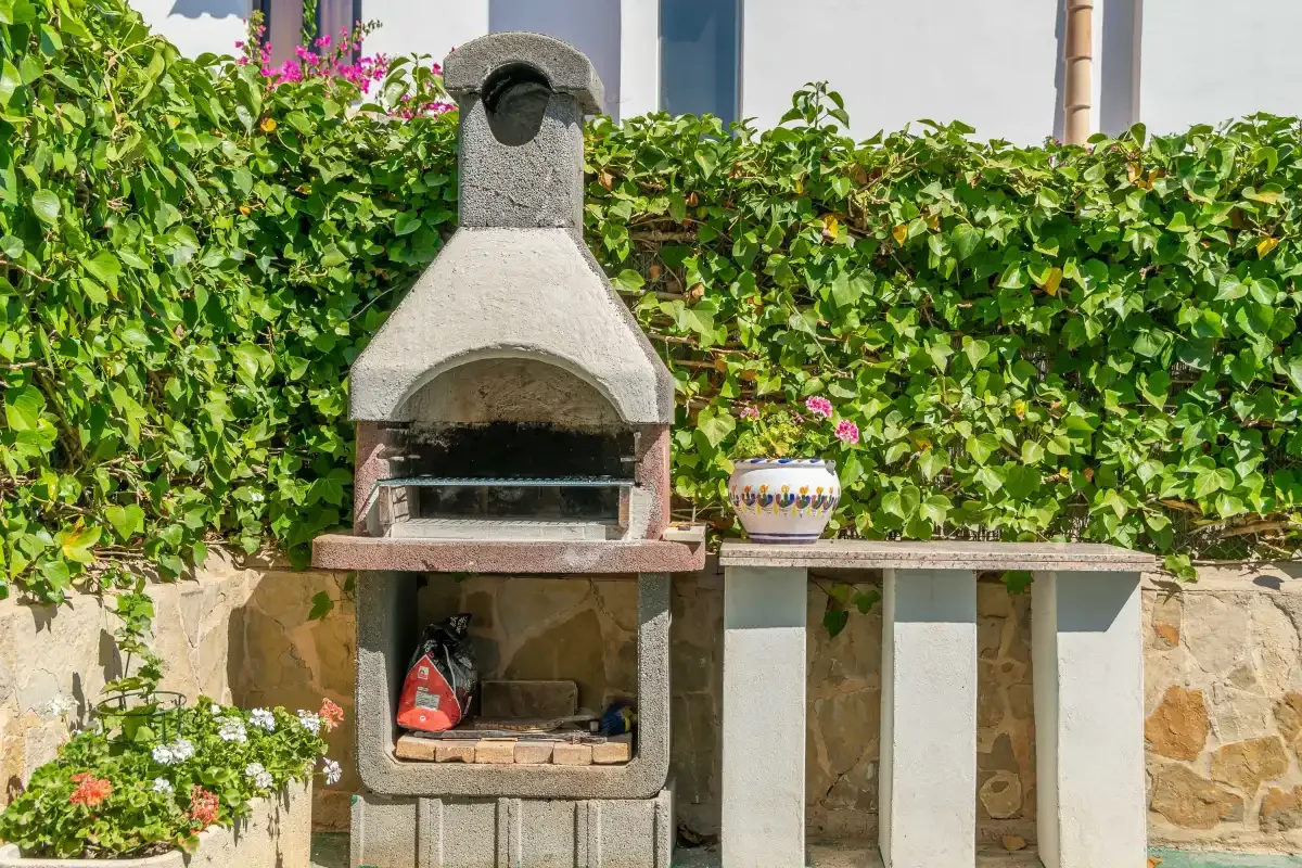 An outdoor barbecue stands beside a tiled counter in a leafy garden.