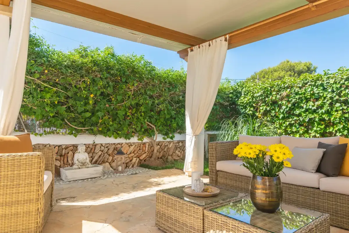 A covered outdoor seating area with curtains, a low table, and green hedges.