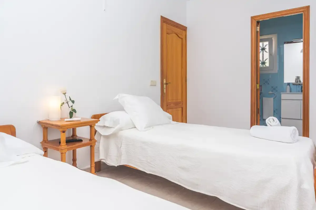 A bright bedroom with white bedding, a wooden bedside table, and an open bathroom doorway.