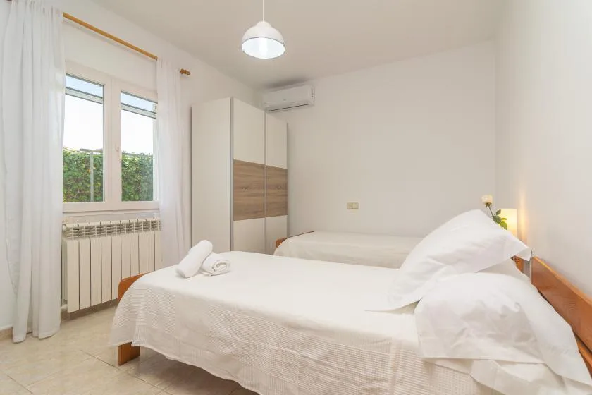 A bright bedroom with two beds, a window, and white bedding.
