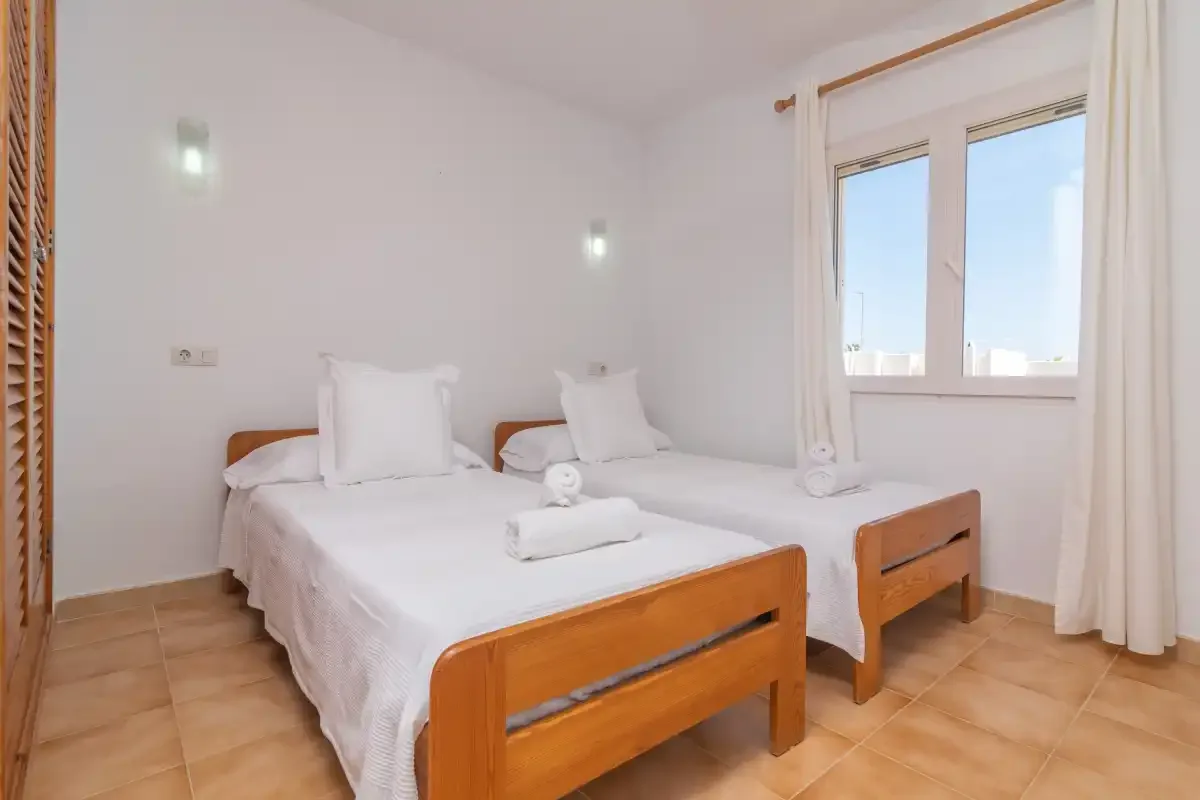 A bright bedroom with two beds and white linens beside a window.
