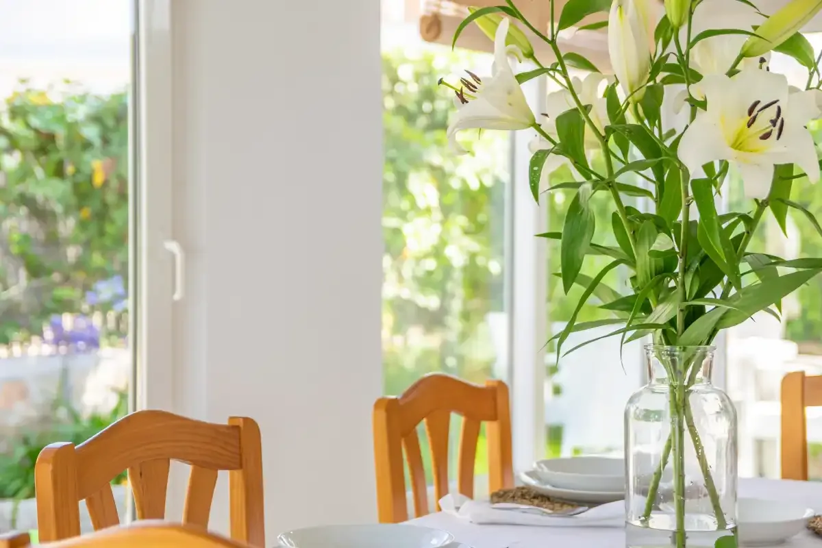 A dining area by bright windows features a vase of white flowers on the table.