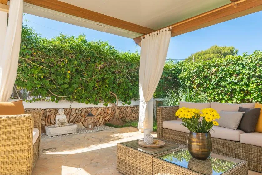 A covered patio seating area with a wicker table and flowering plants.