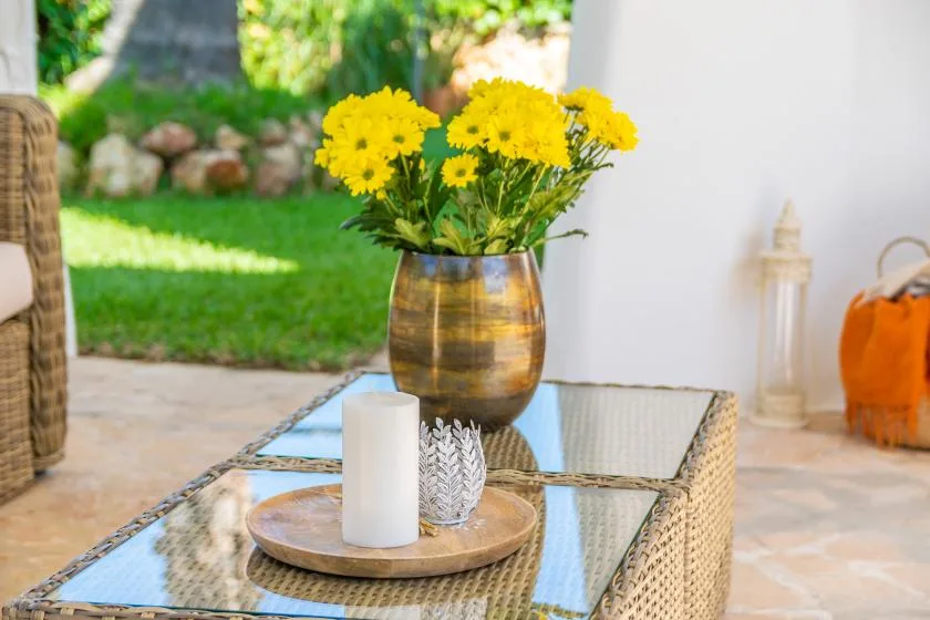 A glass-topped outdoor table holds a vase of yellow flowers and a tray.