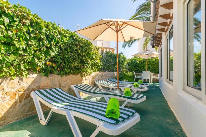 Outdoor poolside seating area with striped loungers, sun umbrellas, and leafy hedges.