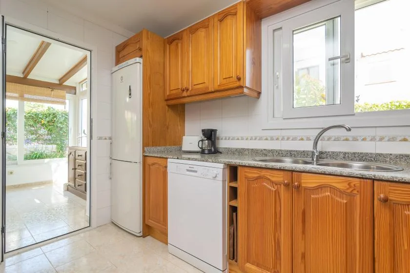 A bright kitchen with wooden cabinets, a refrigerator, and a sink beneath the window.