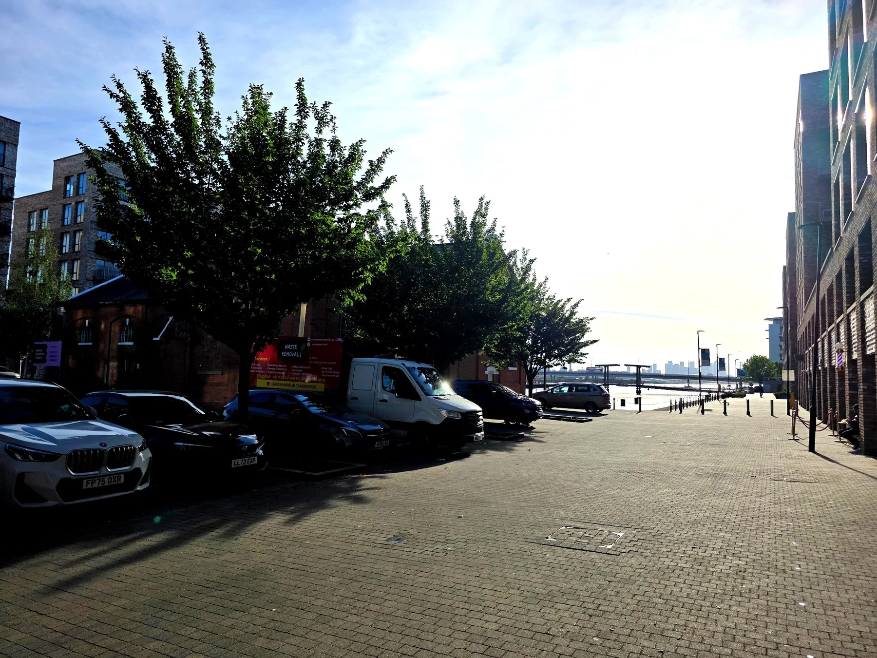 A paved street between buildings, with parked vehicles and trees.