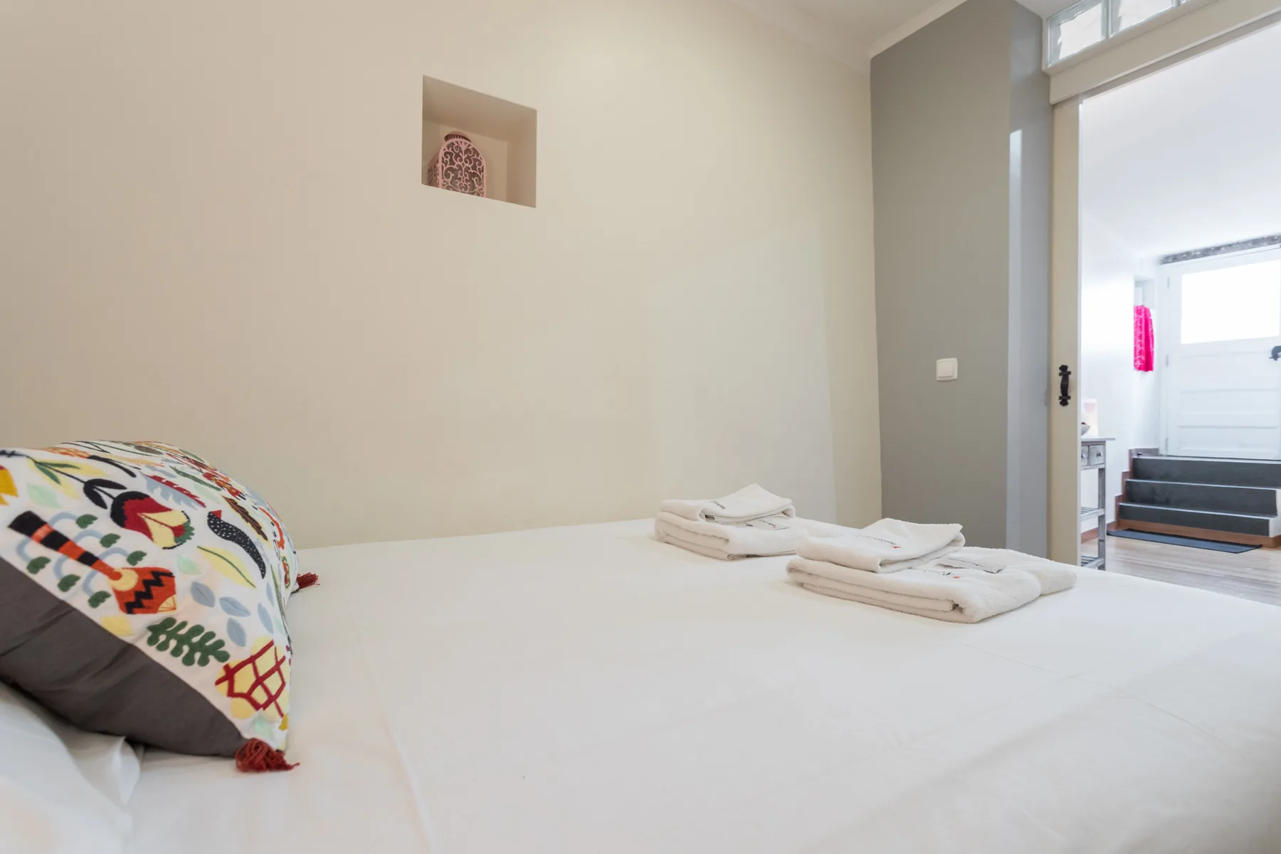 A bedroom with a white bed, folded towels, and a colorful patterned cushion.
