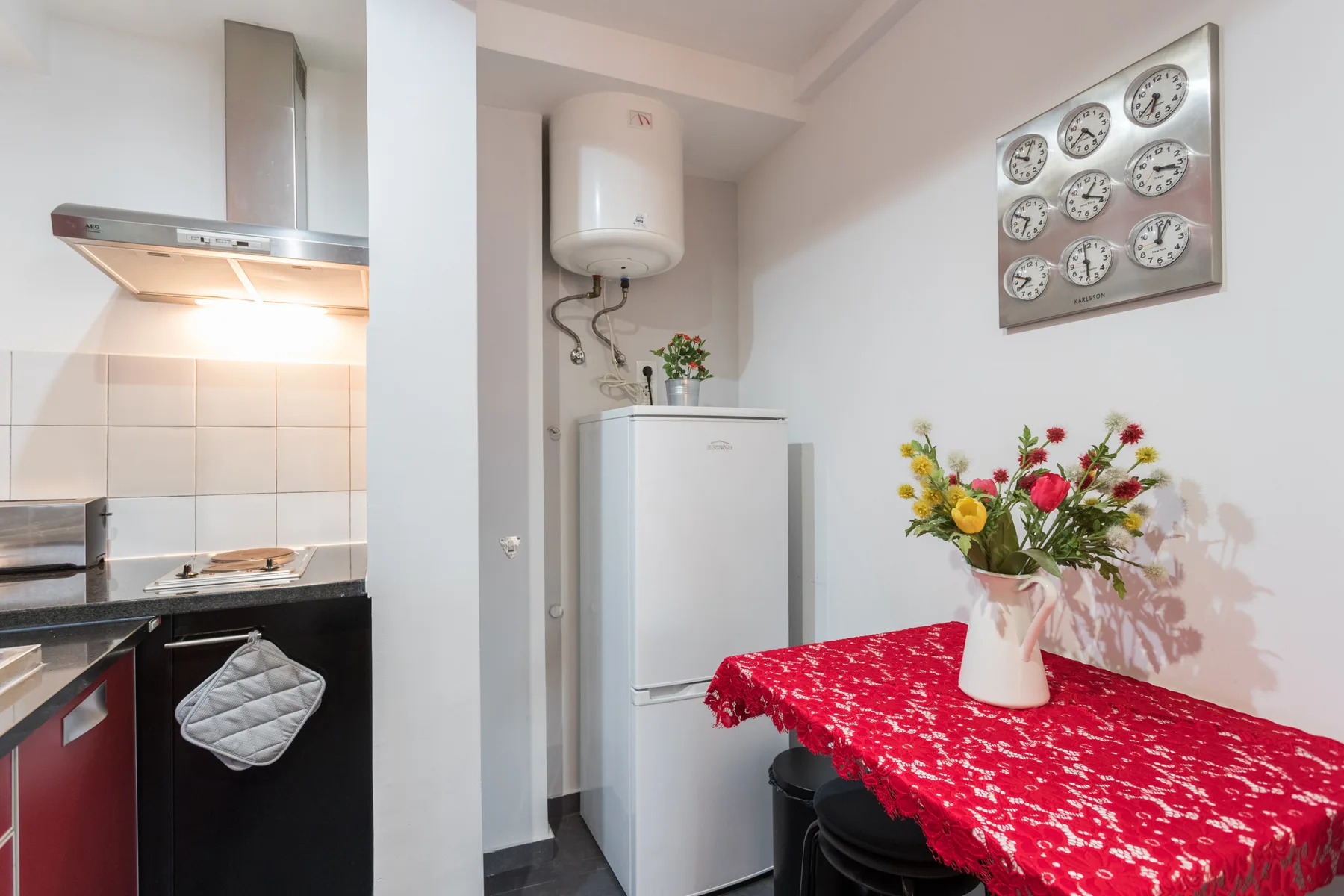 A compact kitchen with a refrigerator and a red-covered table topped with flowers.