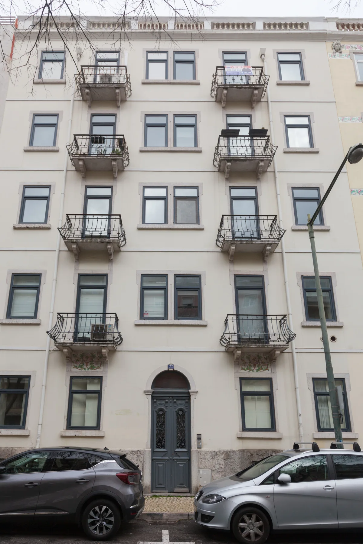 A multi-story apartment building with balconies faces a street with parked cars.