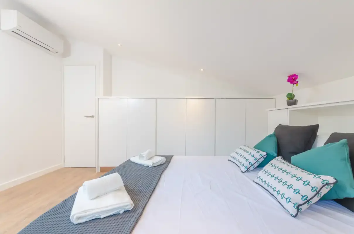 A bright bedroom with a bed, colorful pillows, and a white wardrobe.
