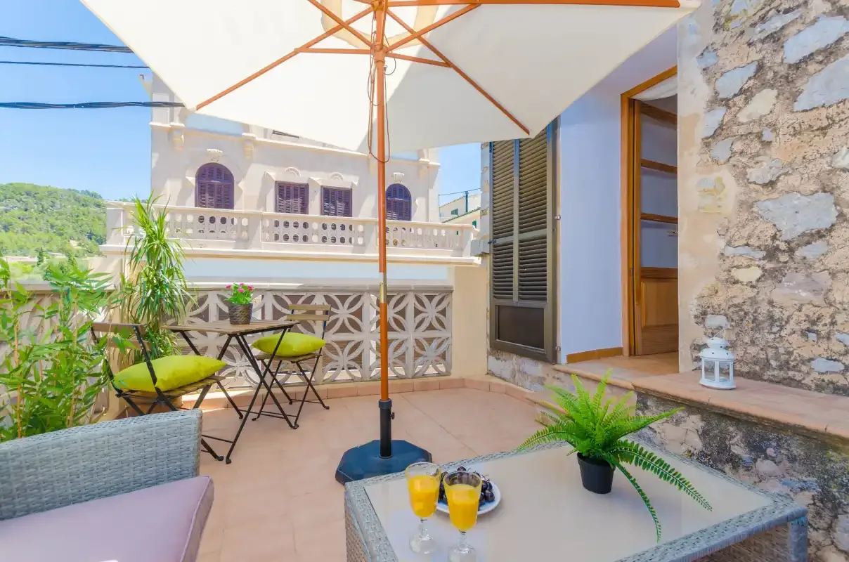 A sunny outdoor patio has a large umbrella, seating, and potted plants.
