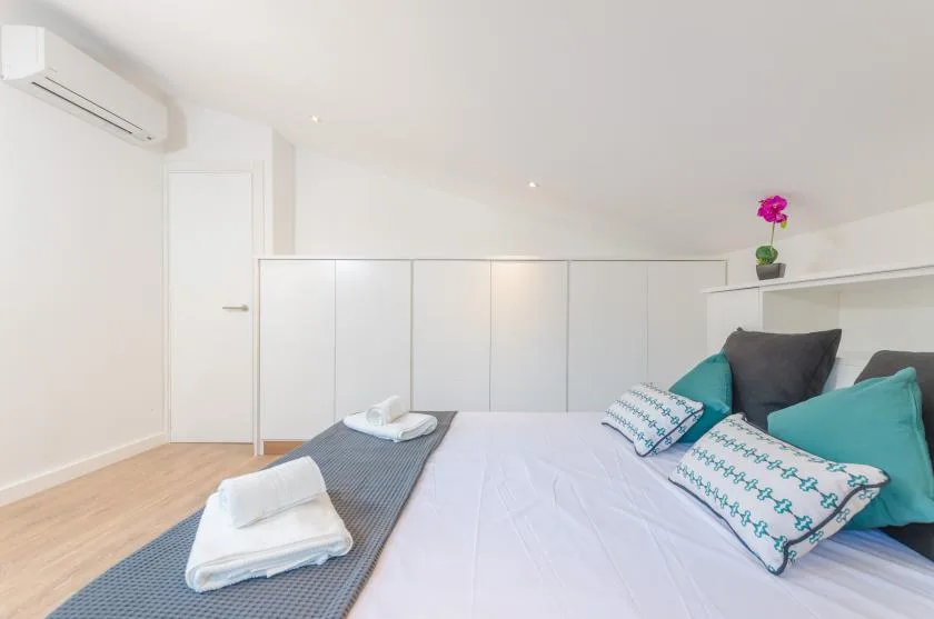 A bright bedroom with a bed, colorful cushions, and a wall-mounted air conditioner.