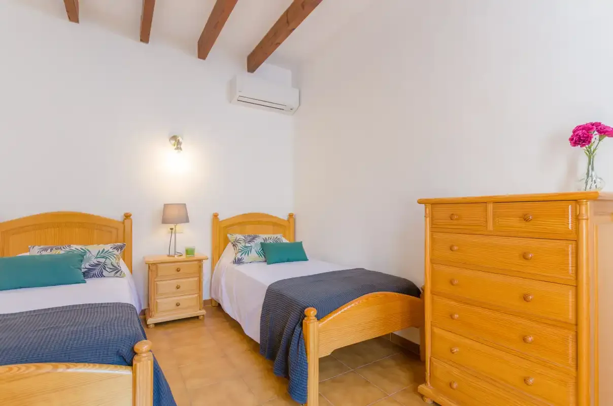 A bright bedroom with two beds and wooden dressers beneath exposed ceiling beams.