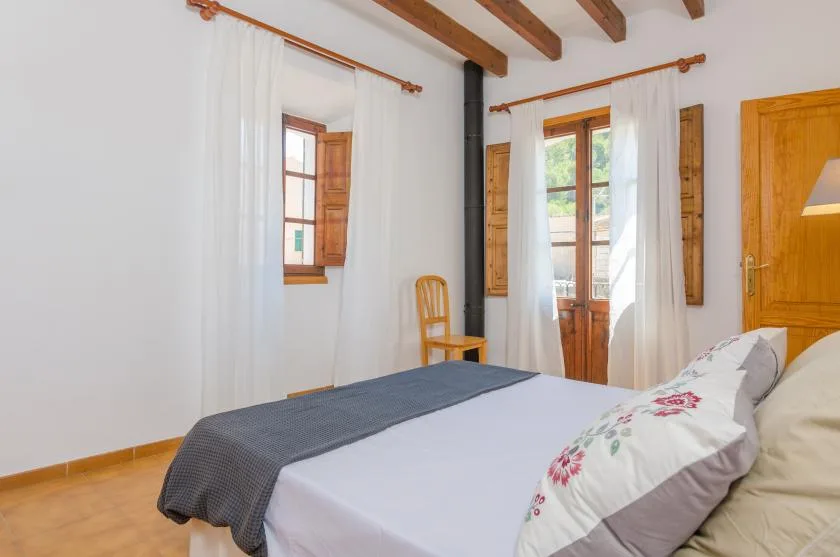 A bedroom with a bed, wooden ceiling beams, and windows draped with white curtains.
