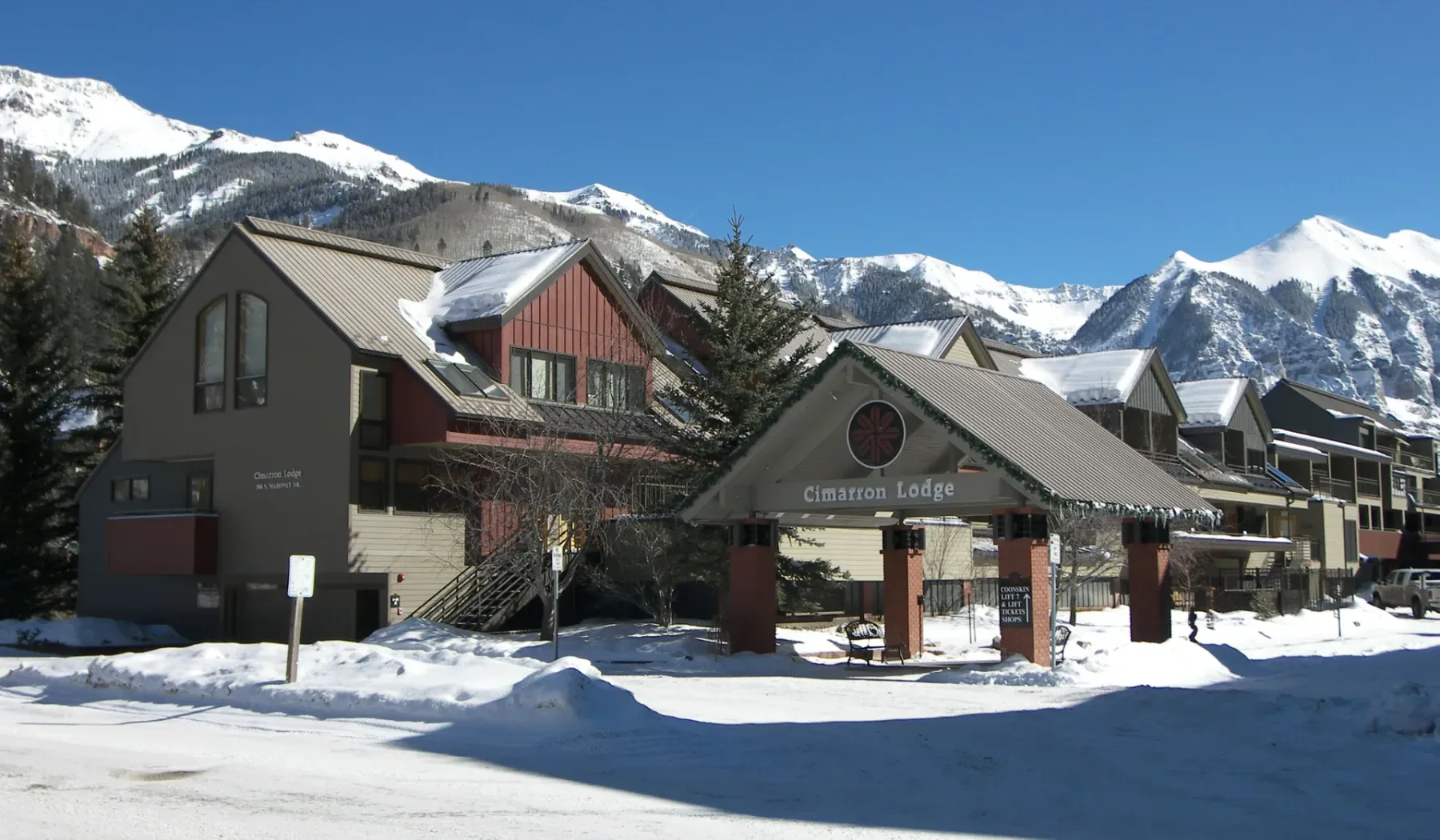 La entrada de un alojamiento aparece junto a edificios nevados, con montañas al fondo.