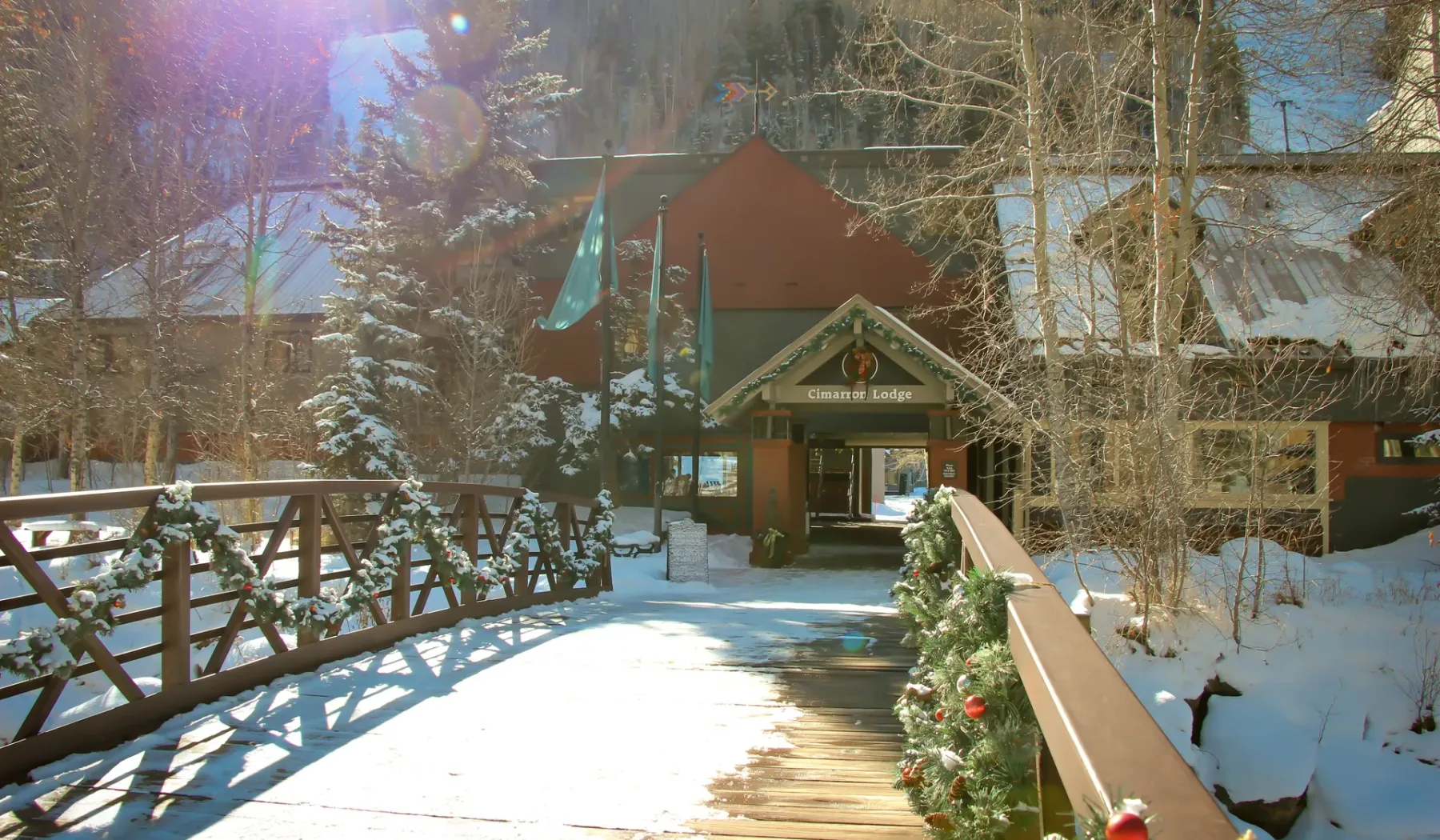 Un camino de madera cubierto de nieve conduce a la entrada de un albergue decorada con vegetación.