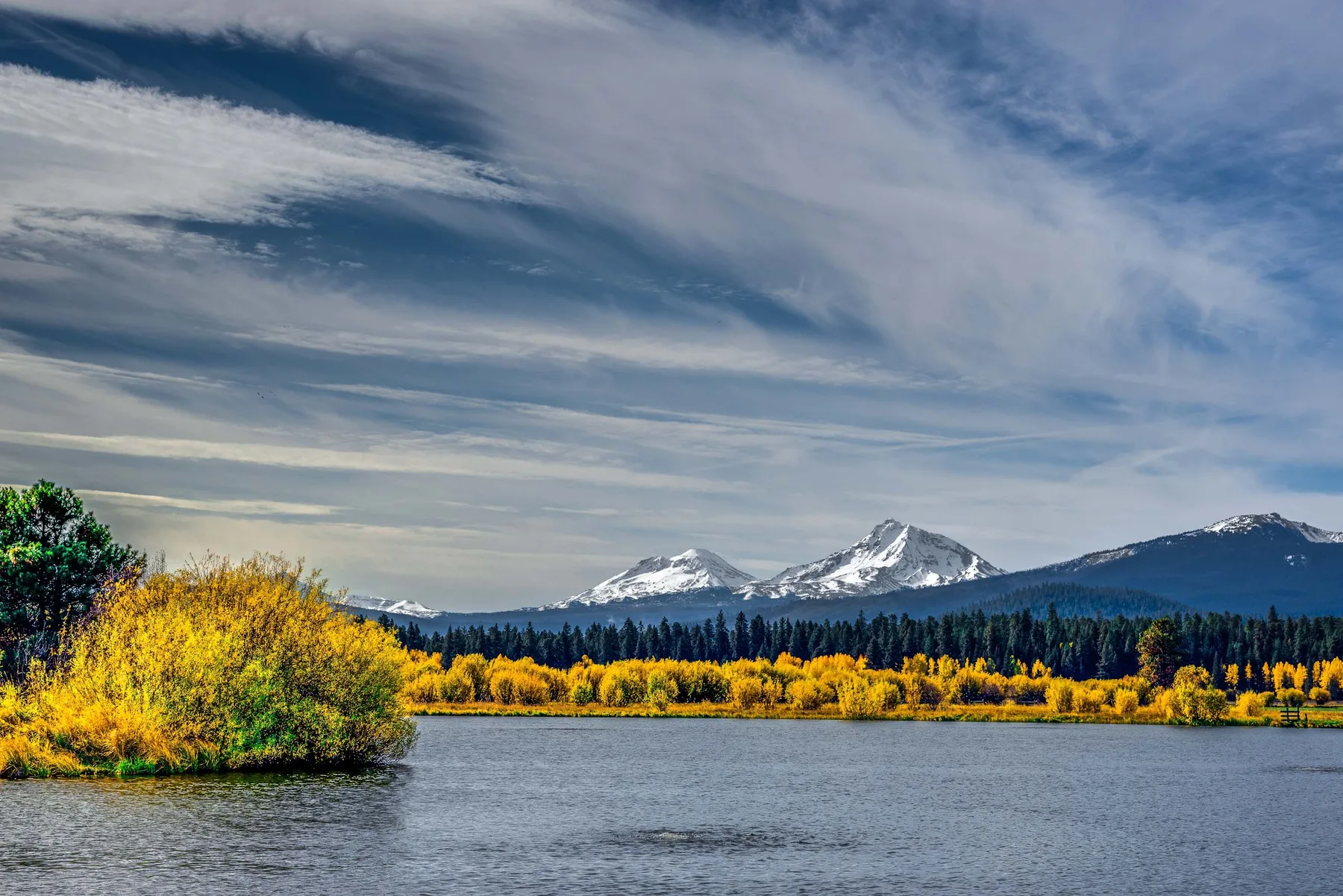A lake stretches toward a line of golden autumn trees and snow-capped mountains.