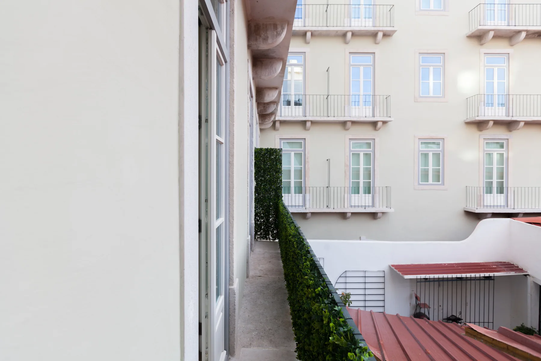A narrow outdoor walkway runs beside a hedge and apartment windows.
