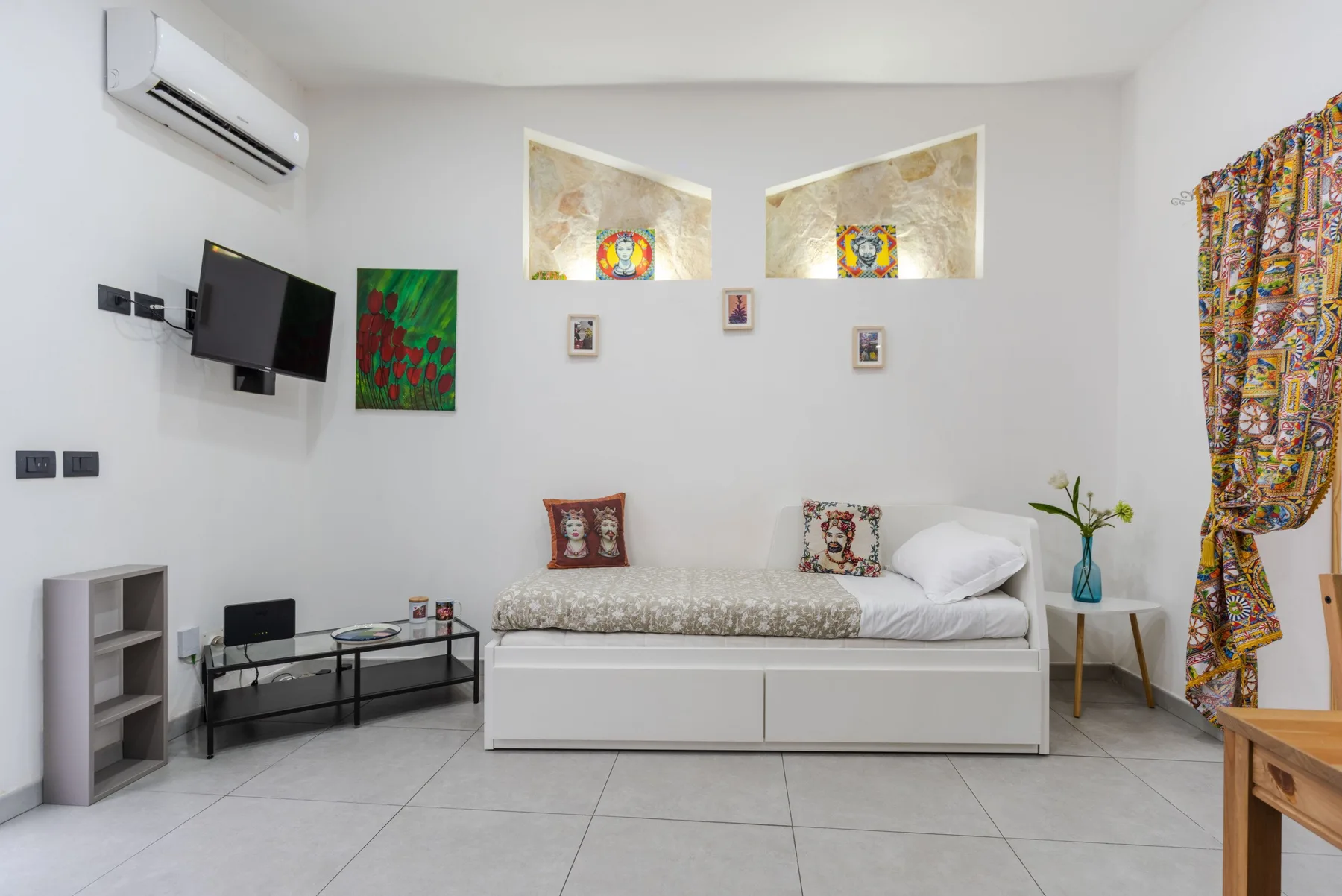 A bright bedroom with a single bed, wall-mounted television, and colorful curtains.