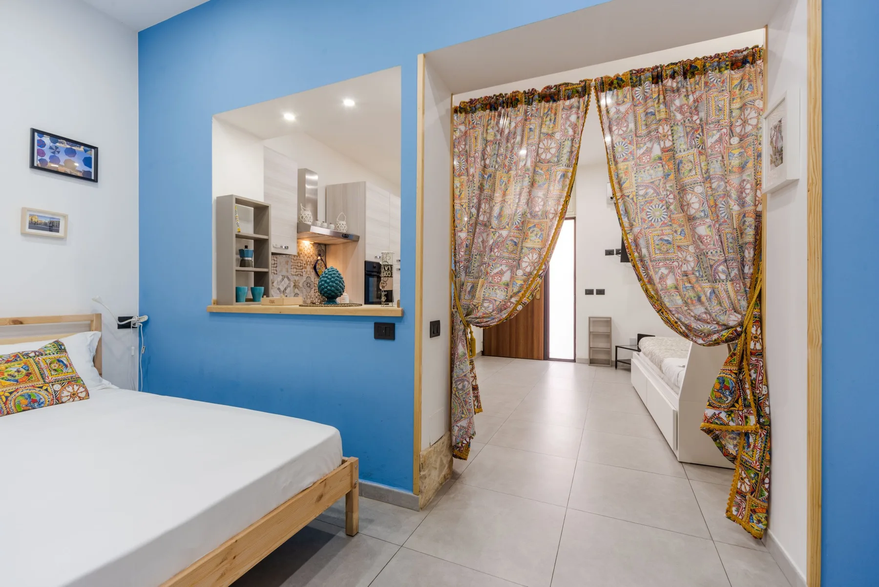 A bedroom opens through a blue wall to a bright adjoining space with patterned curtains.