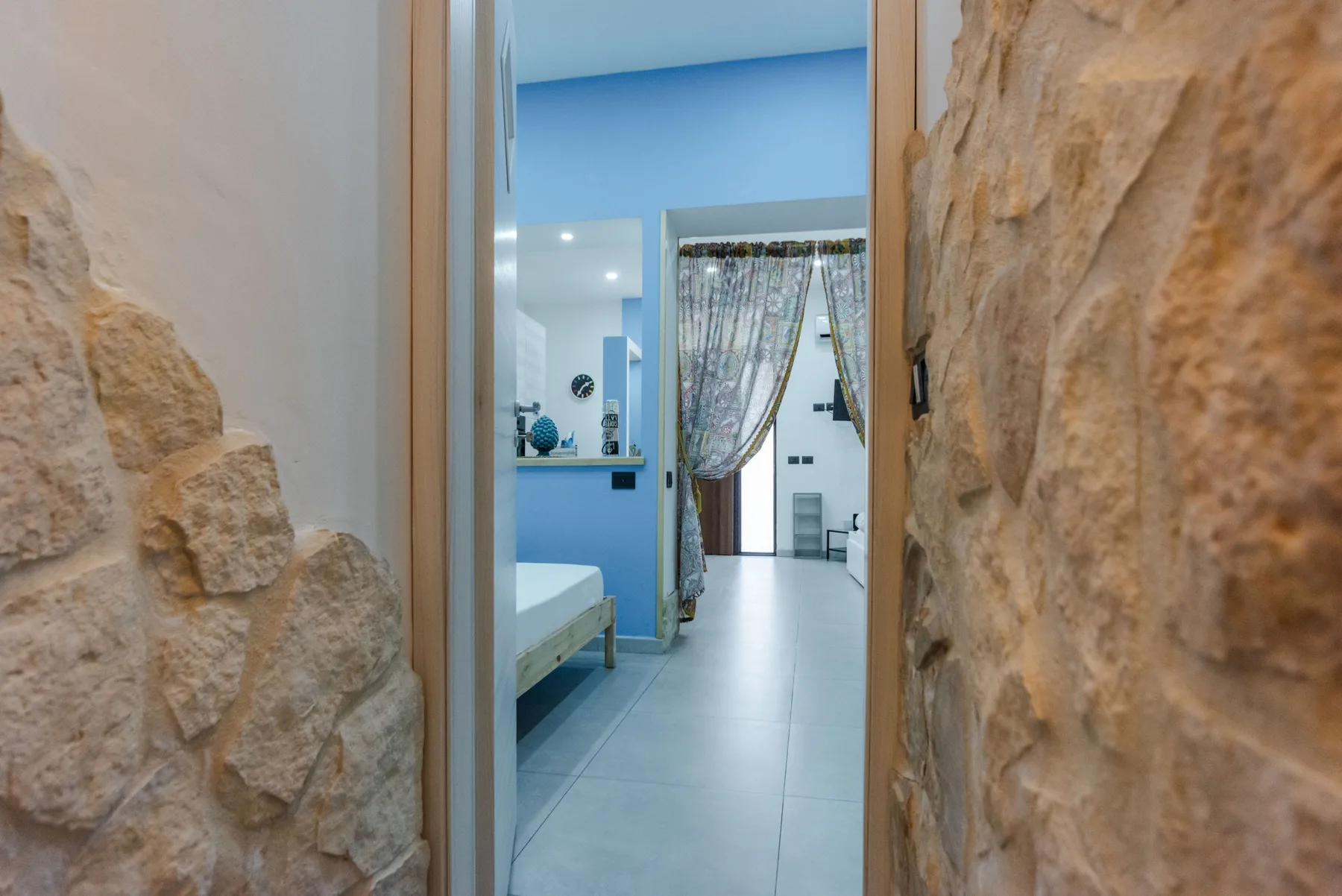A blue bedroom opens onto a bright adjoining space through a curtained doorway.