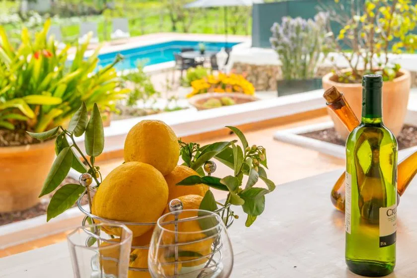 A bottle and glasses sit beside a bowl of lemons on an outdoor table.