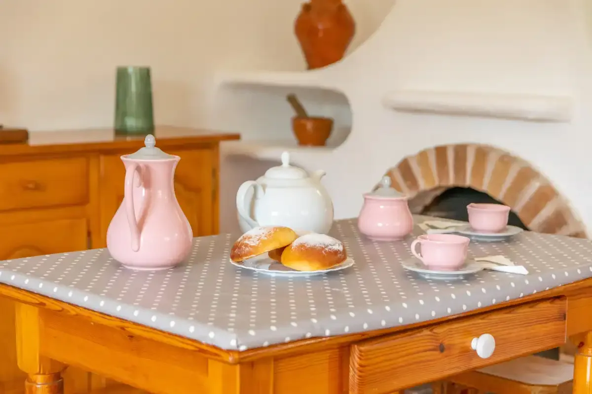 A kitchen table displays teaware and pastries, with a fireplace in the background.