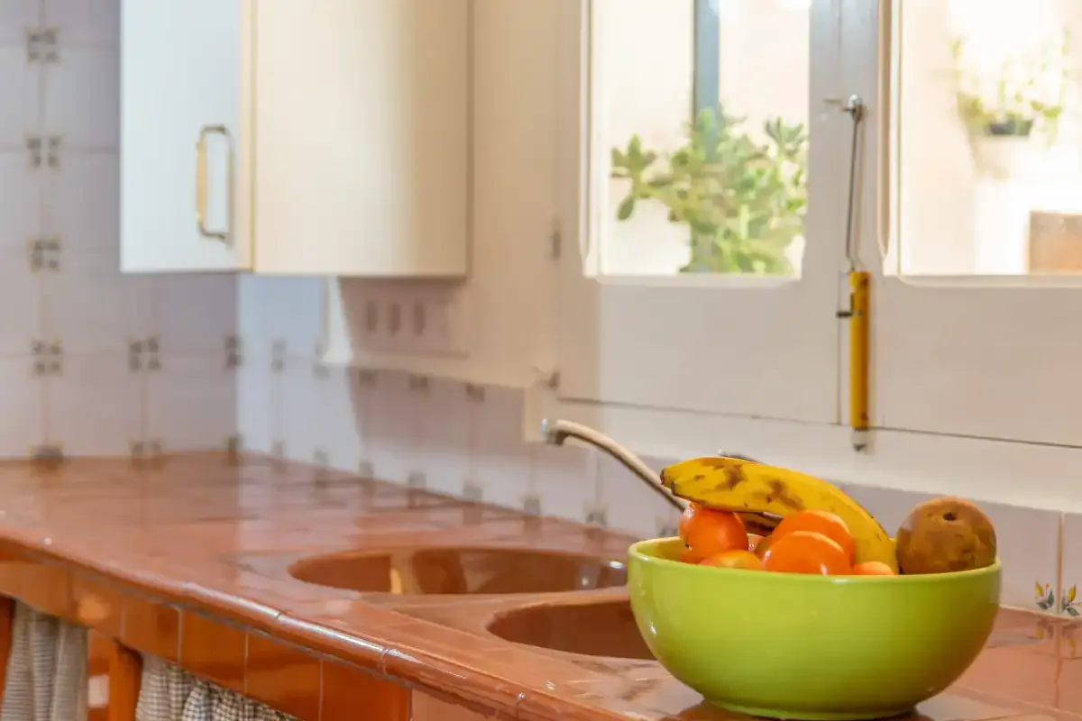 A tiled kitchen counter with a double sink and a bowl of fruit.