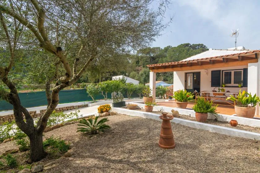 A house opens onto a sunny garden patio with potted plants and trees.