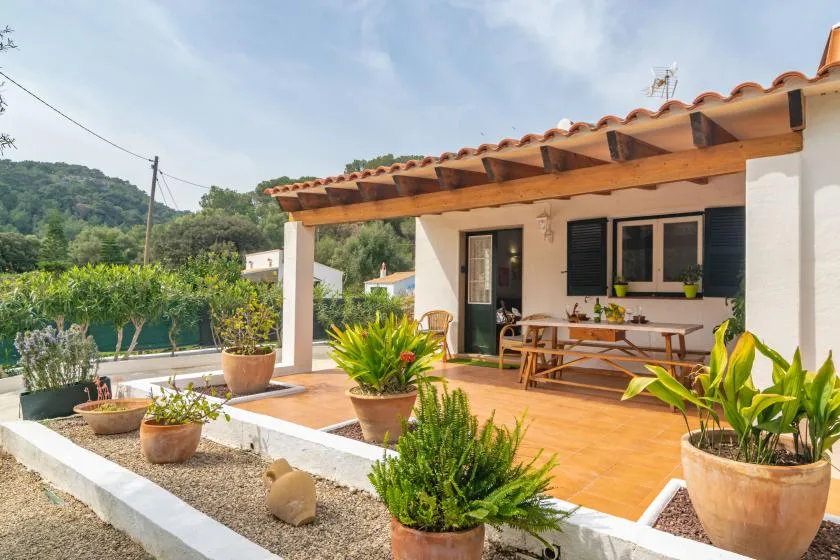 A tiled patio beside a house has outdoor seating and potted plants under a wooden pergola.