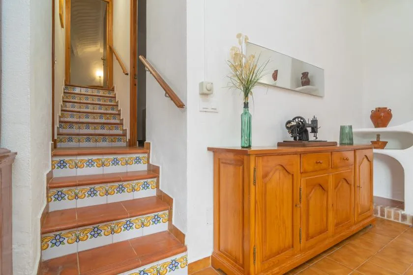 A tiled staircase rises beside a wooden sideboard in a bright interior.