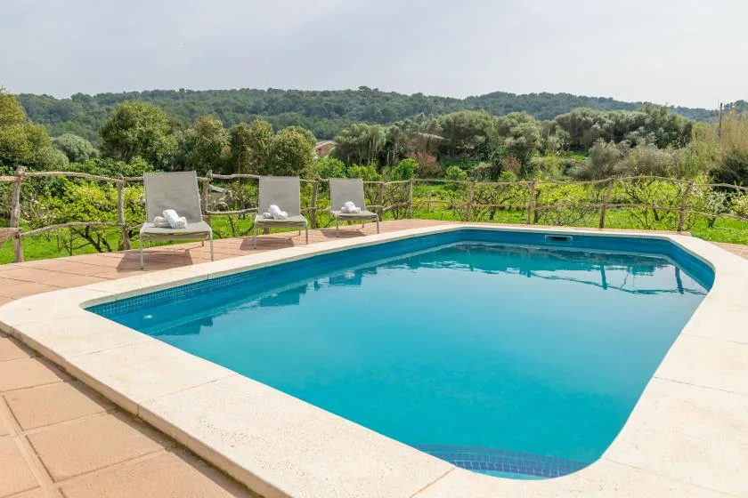 An outdoor swimming pool is surrounded by a paved deck and three lounge chairs.