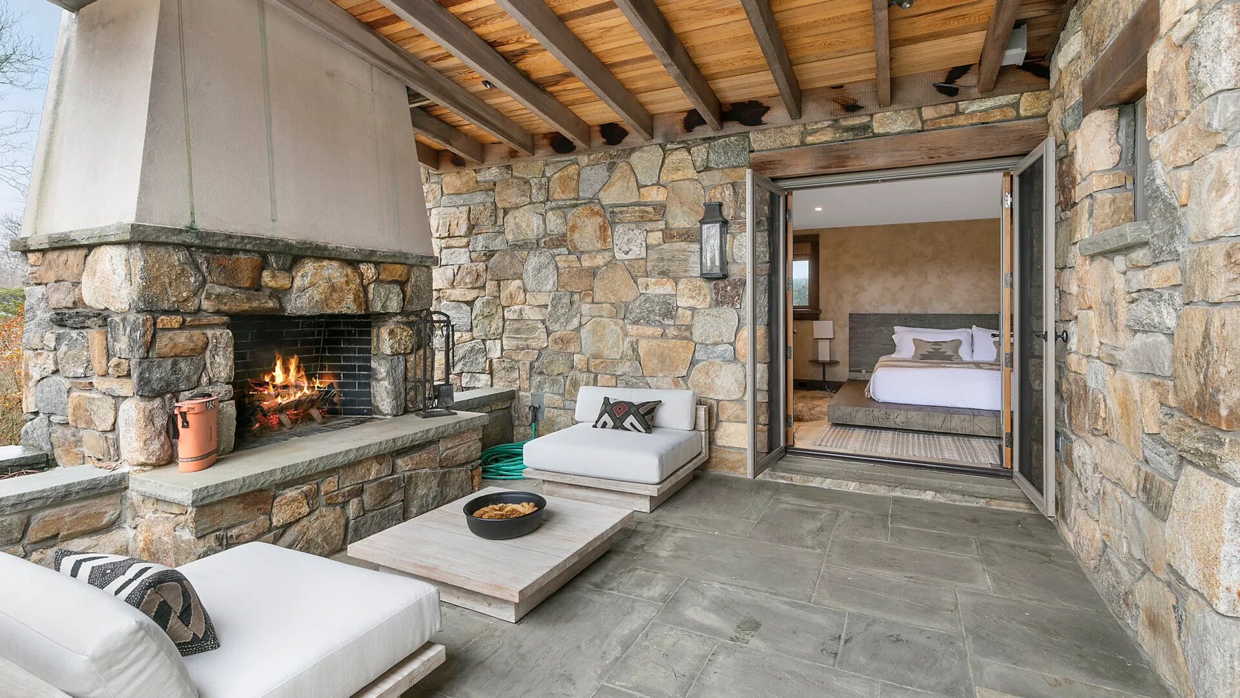 Un patio de piedra con chimenea y asientos junto a una puerta abierta al dormitorio.
