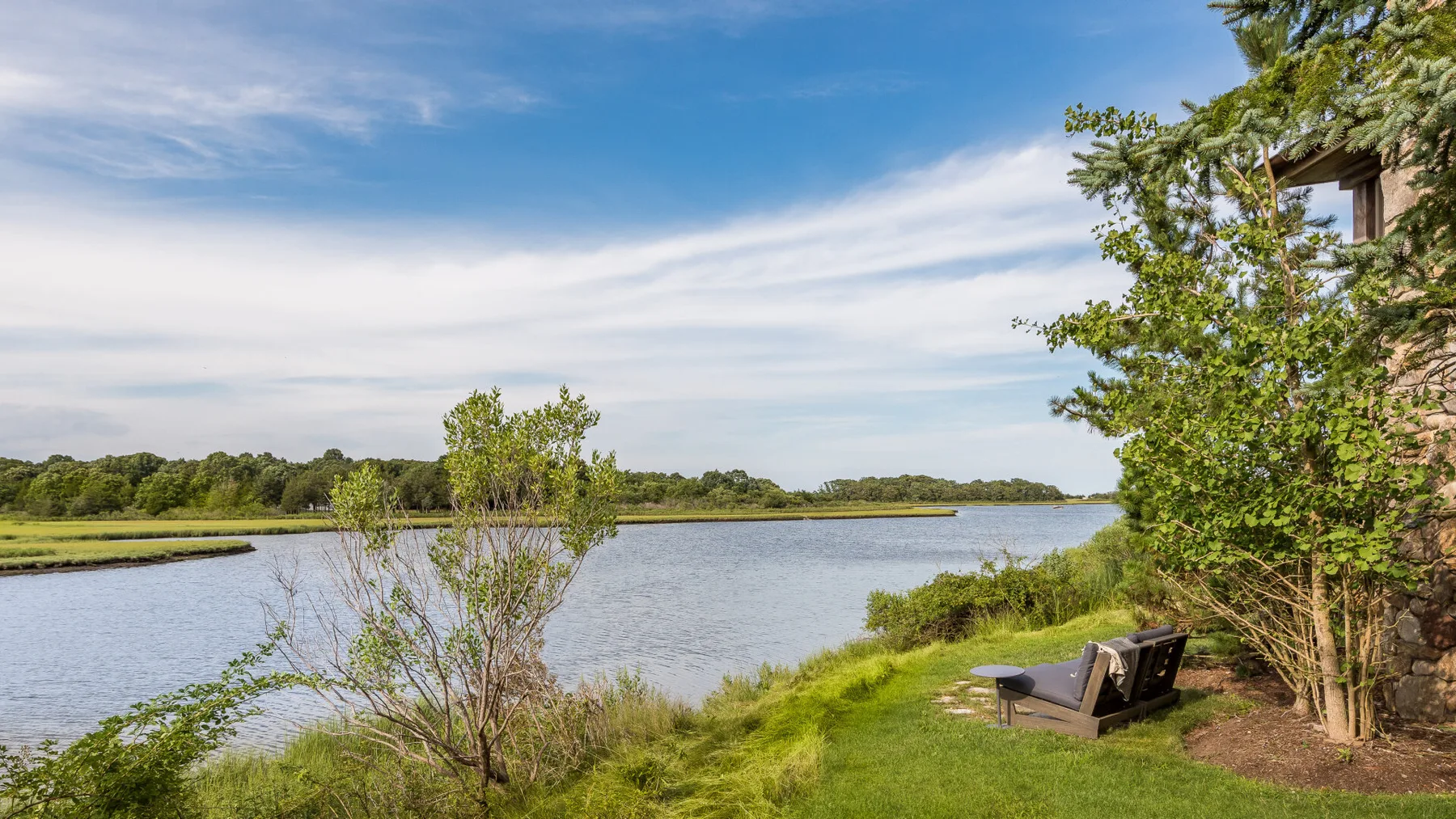 Una orilla cubierta de hierba junto a un lago, con asientos al aire libre y árboles bajo un cielo amplio.