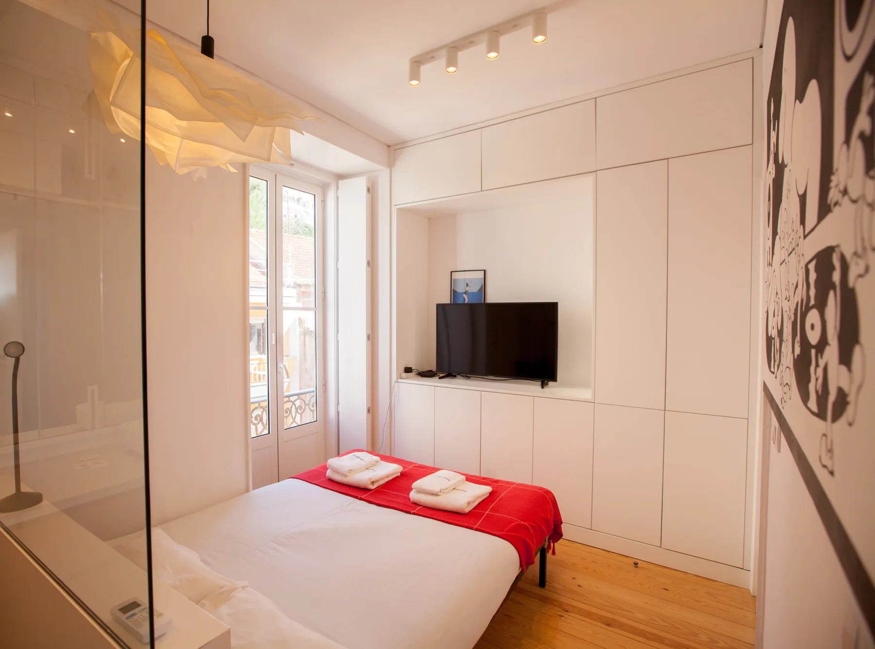 Dormitorio con una cama vestida de blanco y rojo y un televisor en la pared.