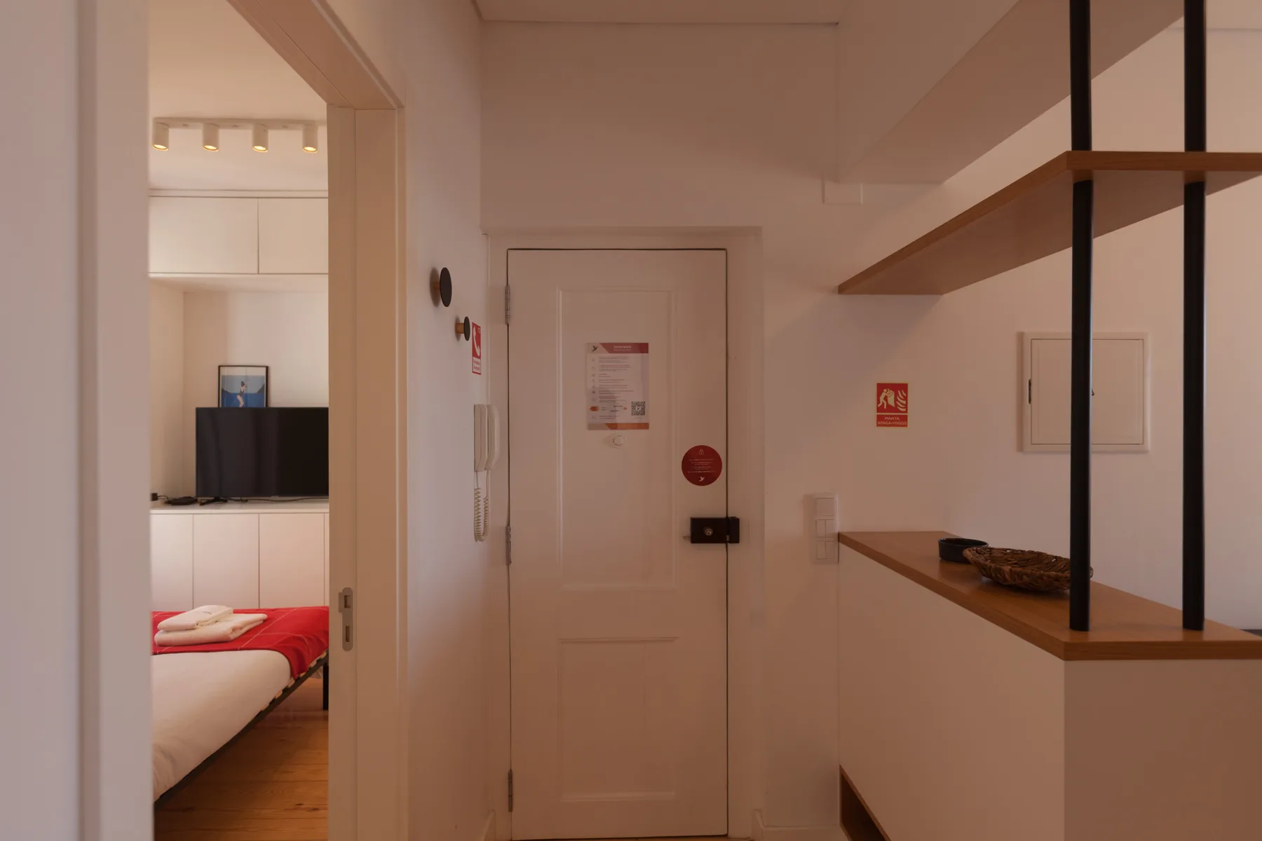 Un pasillo estrecho con una puerta cerrada y un dormitorio visible a través de una puerta abierta.