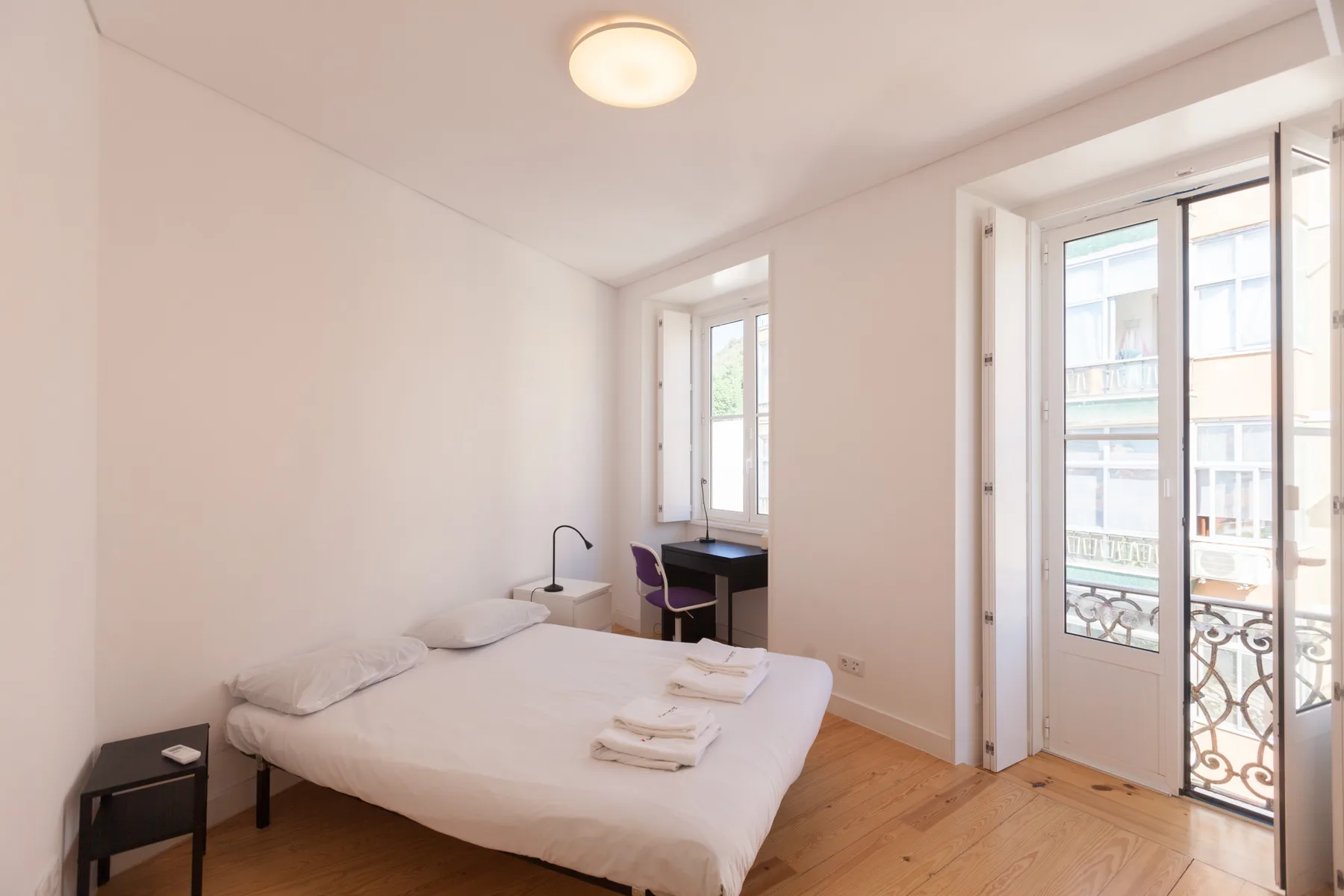 Un dormitorio luminoso con una cama, un escritorio y ventanas altas que dan a un balcón.