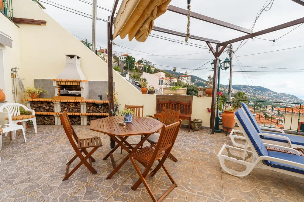 Una terraza al aire libre tiene una mesa con sillas de madera, tumbonas y una parrilla.