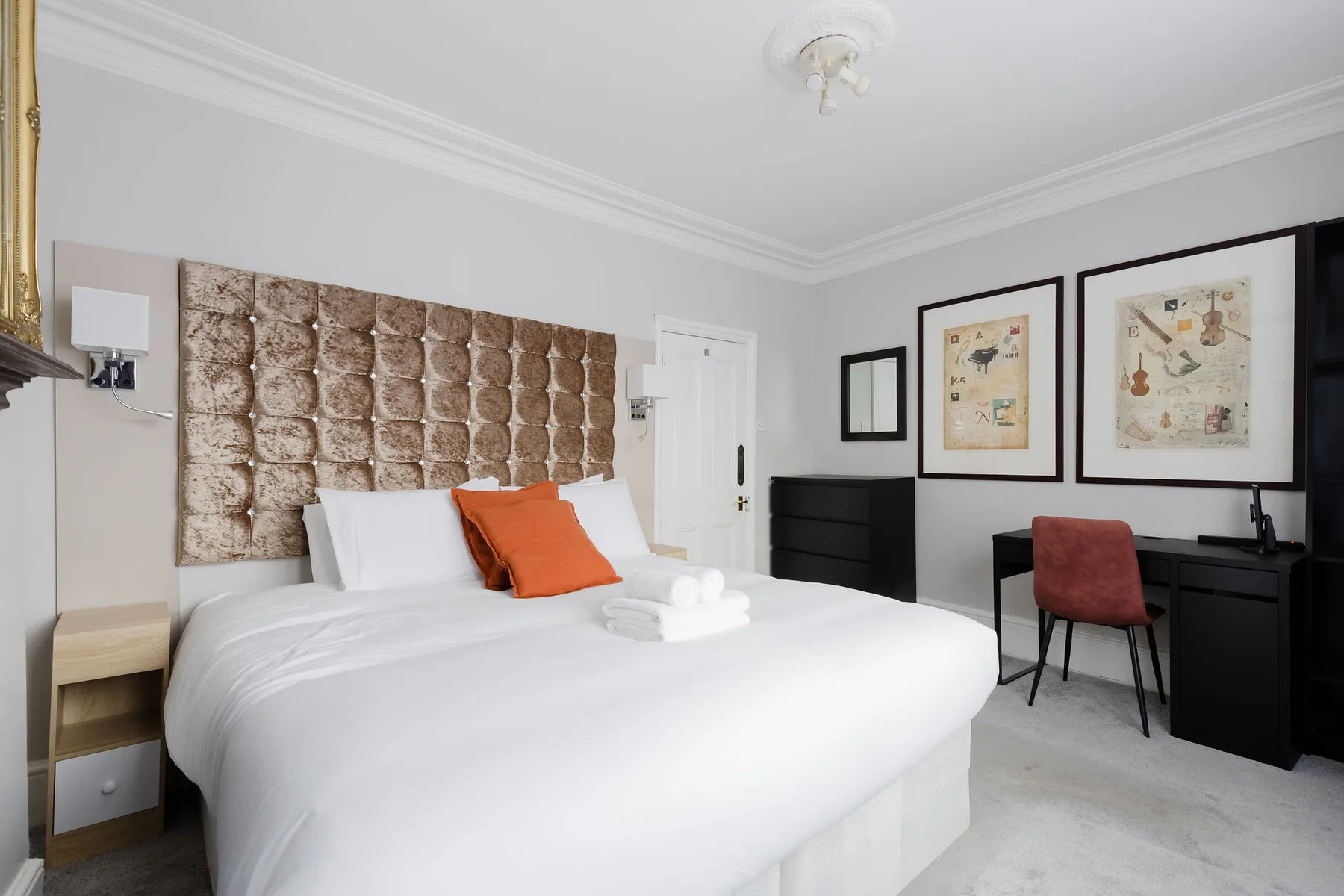 Un dormitorio luminoso con una cama blanca, un cojín naranja y un escritorio negro.